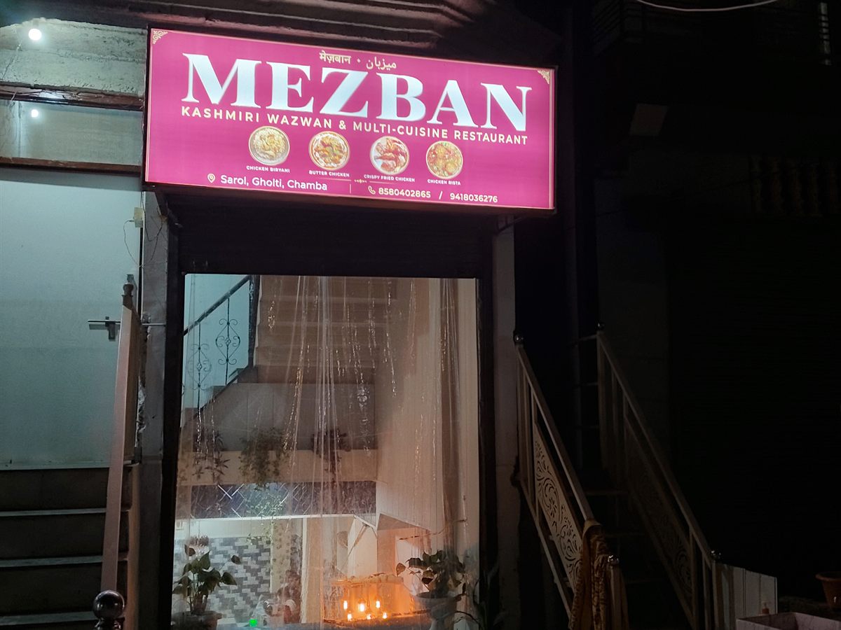 Mezban's lit pink signboard at night above the entrance, reading Kashmiri Wazwan and Multi-Cuisine Restaurant, Sarol Gholti, Chamba.