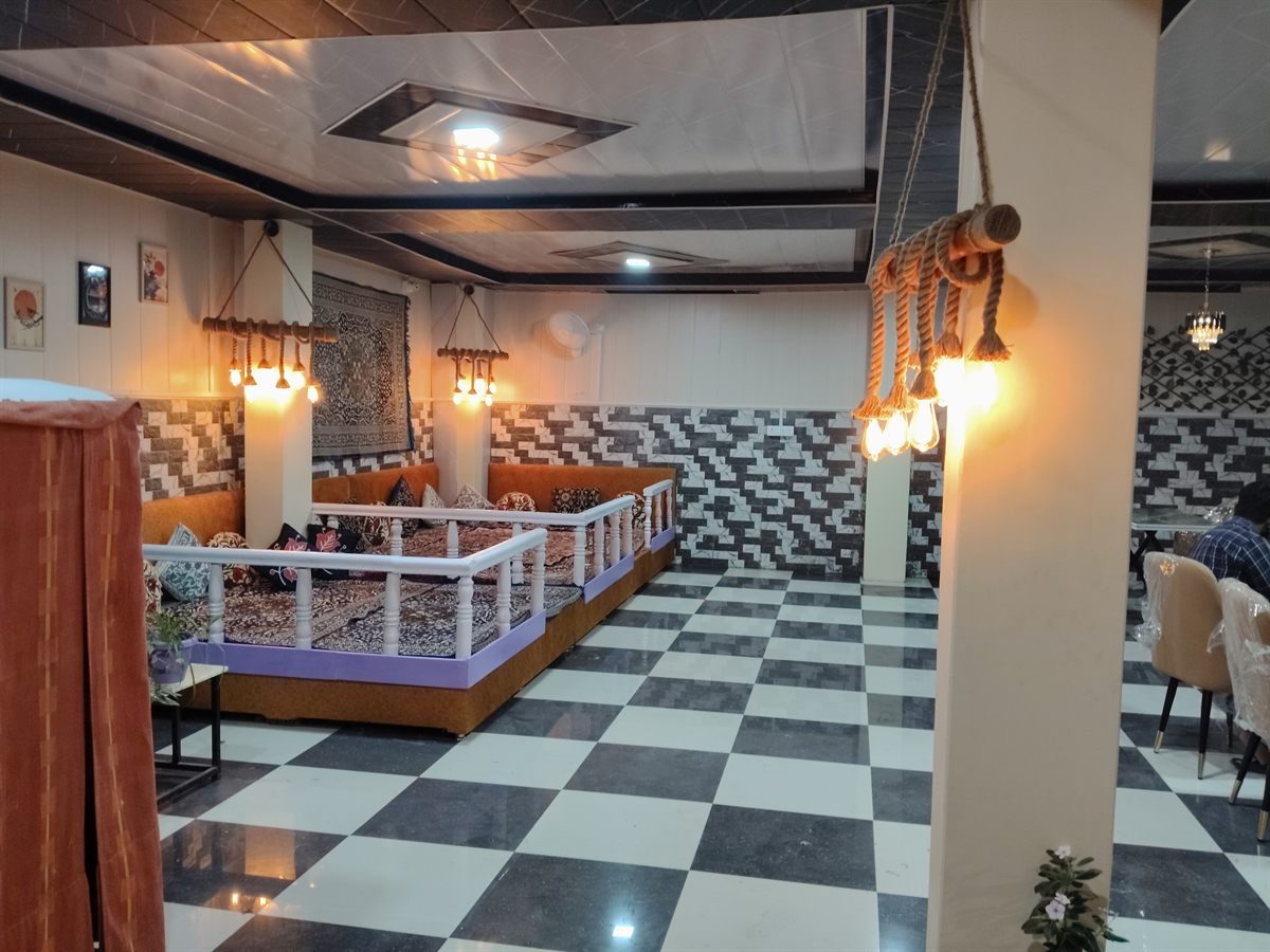 A raised floor-seating platform with orange banquettes, patterned cushions and Kashmiri carpets, lit by rope-hung filament lamps, beside a black and white checkerboard marble floor.