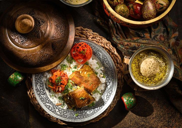 An engraved silver plate of rice topped with fried fish and tomato, set on a woven mat beside a carved copper lidded pot, a bowl of green chutney and painted Kashmiri papier-mâché beads.